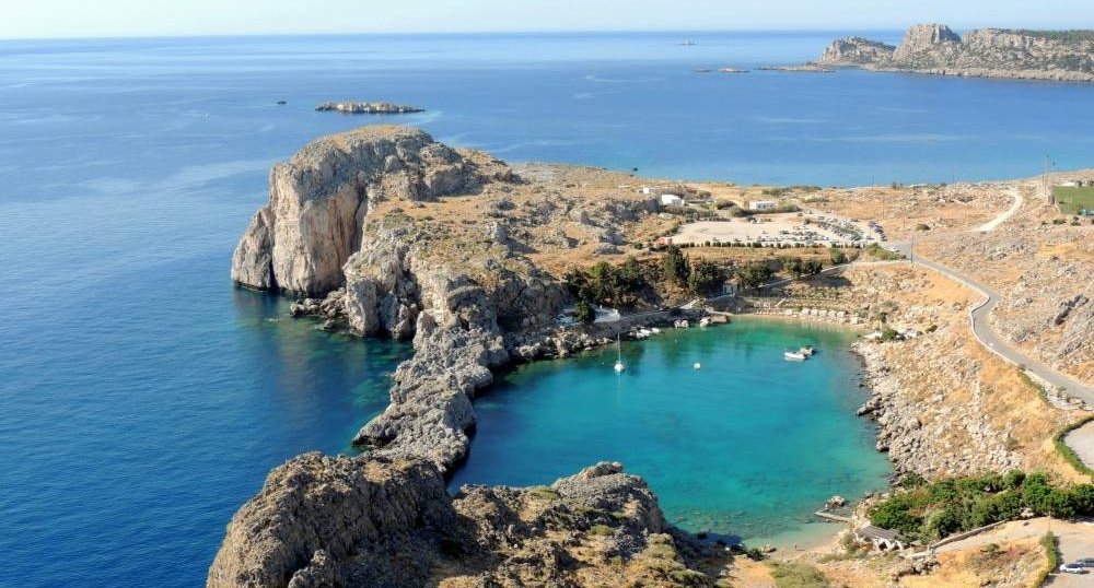 A stunning aerial view of St. Paul's Bay in Lindos, Rhodes, Greece. This picturesque cove, surrounded by dramatic cliffs and turquoise waters, is a popular destination for swimming, snorkeling, and relaxation.